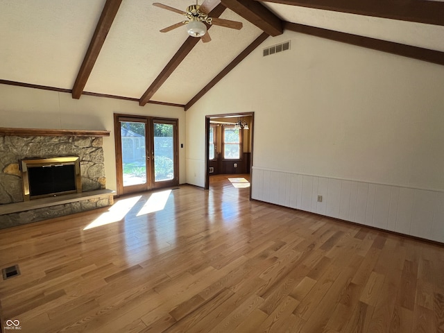 unfurnished living room with ceiling fan, beam ceiling, french doors, a stone fireplace, and hardwood / wood-style flooring