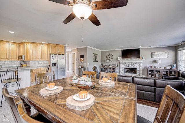 dining room featuring light tile patterned floors, a ceiling fan, crown molding, a stone fireplace, and recessed lighting