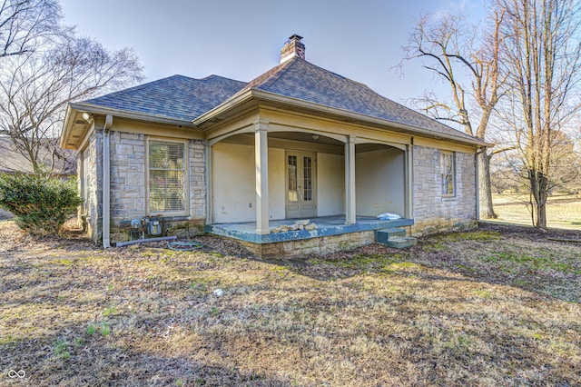 rear view of house with covered porch, stone siding, a shingled roof, and a chimney