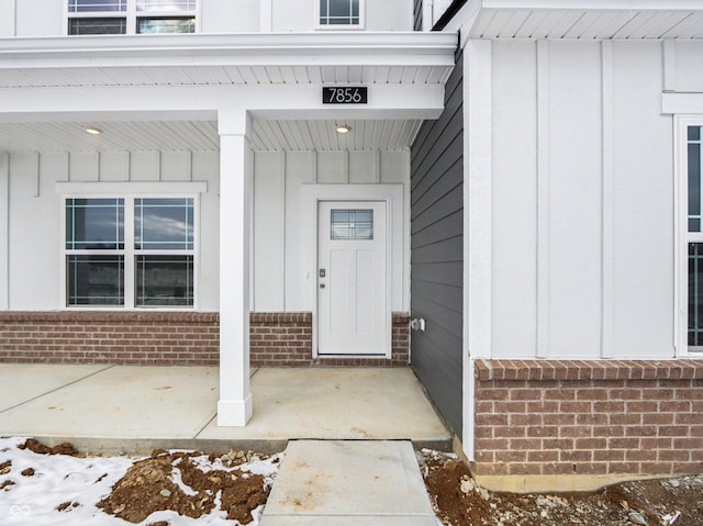 snow covered property entrance with covered porch, board and batten siding, and brick siding