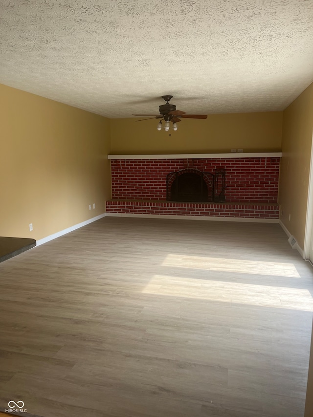 empty room with a brick fireplace, ceiling fan, wood-type flooring, and a textured ceiling