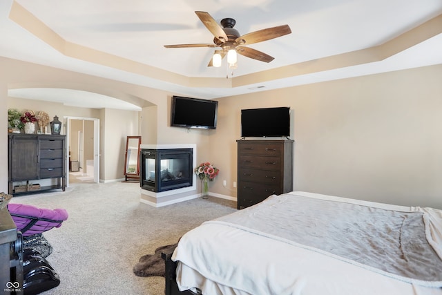 carpeted bedroom with a multi sided fireplace, a tray ceiling, and ceiling fan