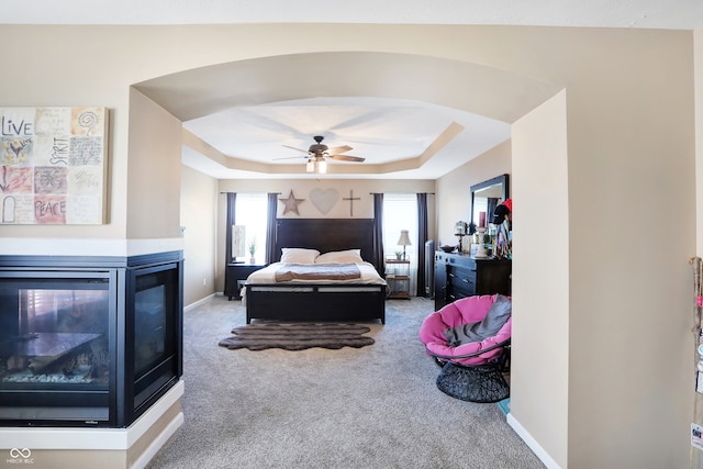 carpeted bedroom featuring a multi sided fireplace, ceiling fan, and a raised ceiling