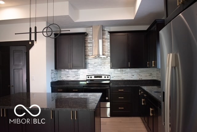 kitchen featuring wall chimney exhaust hood, appliances with stainless steel finishes, dark stone counters, and pendant lighting