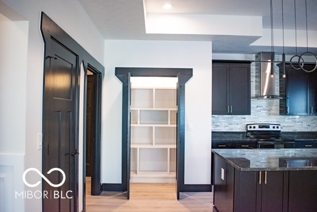 kitchen featuring light wood-type flooring, backsplash, stainless steel range with electric stovetop, hanging light fixtures, and wall chimney exhaust hood