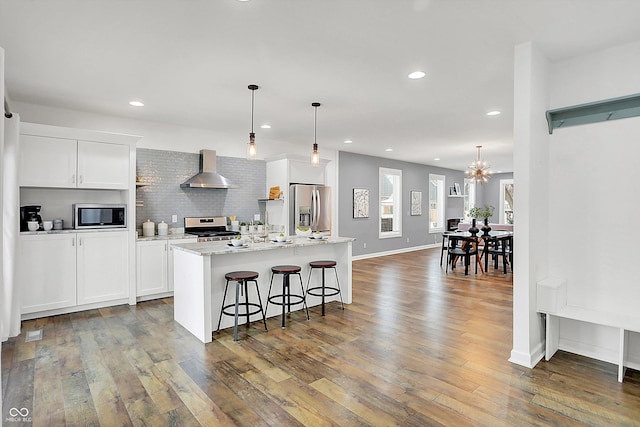 kitchen featuring appliances with stainless steel finishes, wall chimney exhaust hood, decorative light fixtures, white cabinetry, and a center island with sink