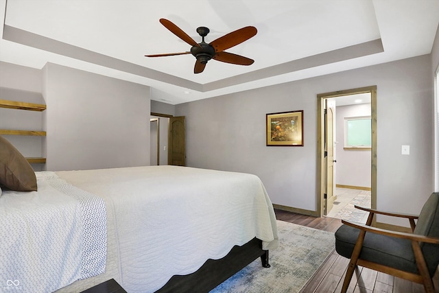 bedroom with ensuite bathroom, a tray ceiling, light wood-type flooring, and ceiling fan