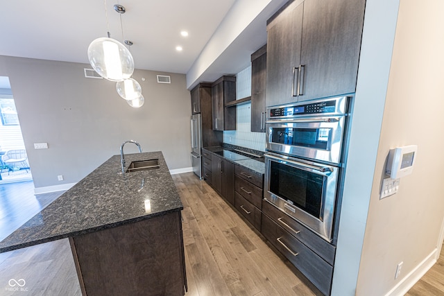 kitchen with a kitchen island with sink, dark brown cabinets, stainless steel appliances, dark stone countertops, and light hardwood / wood-style floors