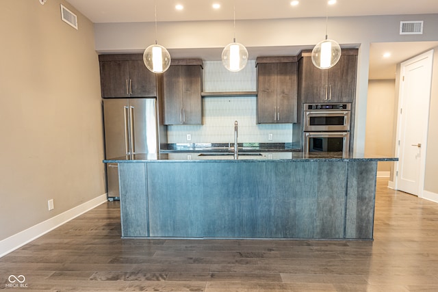 kitchen with appliances with stainless steel finishes, dark brown cabinets, dark stone countertops, dark hardwood / wood-style floors, and a kitchen island with sink