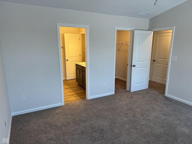 unfurnished bedroom featuring ensuite bath, a spacious closet, dark colored carpet, vaulted ceiling, and a closet