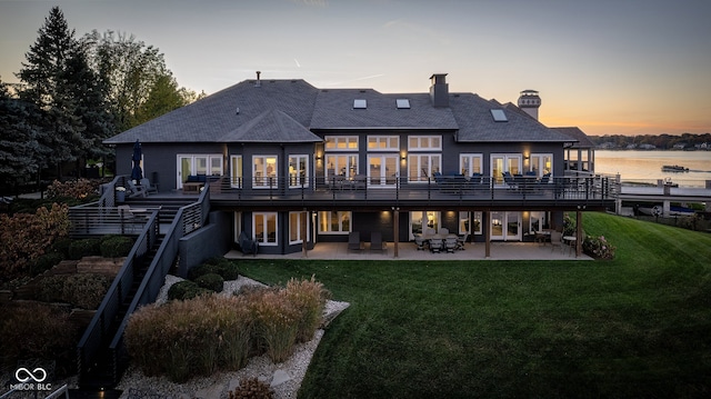 back house at dusk with a water view, a patio area, and a lawn