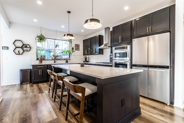 kitchen featuring wall chimney exhaust hood, hanging light fixtures, stainless steel appliances, a center island, and light wood-type flooring