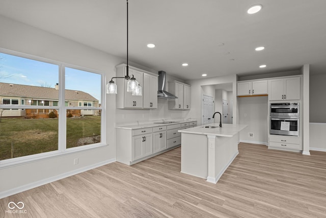 kitchen with wall chimney exhaust hood, stainless steel double oven, decorative light fixtures, black electric cooktop, and a center island with sink