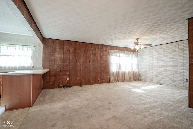 carpeted empty room with wood walls, brick wall, ceiling fan, and plenty of natural light