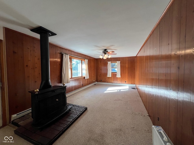 unfurnished living room featuring a baseboard radiator, a ceiling fan, wooden walls, a wood stove, and carpet flooring