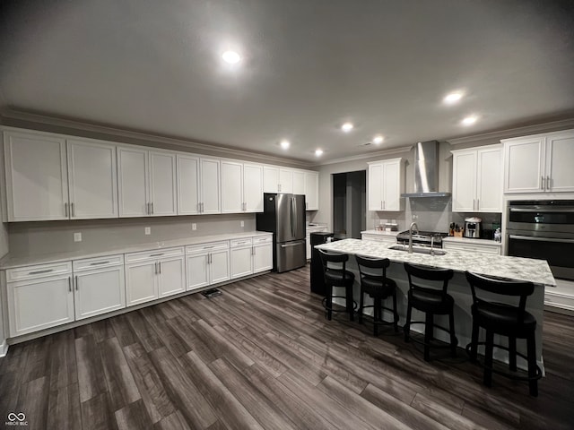 kitchen with a kitchen island with sink, wall chimney exhaust hood, a breakfast bar, white cabinetry, and appliances with stainless steel finishes