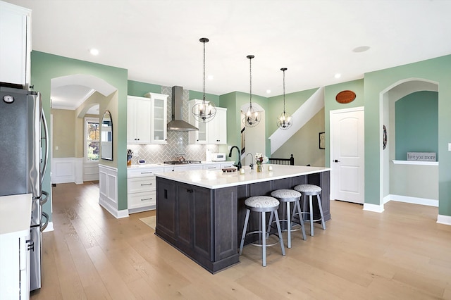 kitchen featuring stainless steel appliances, a center island with sink, white cabinets, wall chimney exhaust hood, and decorative light fixtures