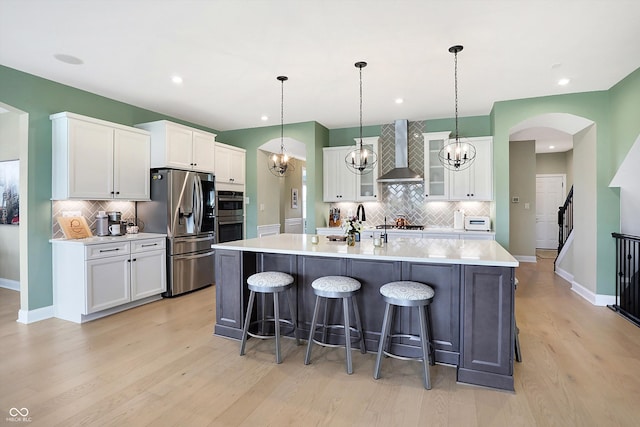 kitchen with stainless steel appliances, light wood-type flooring, wall chimney range hood, an island with sink, and white cabinets