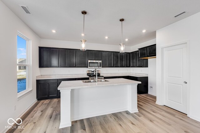 kitchen featuring sink, light hardwood / wood-style flooring, appliances with stainless steel finishes, a center island with sink, and decorative light fixtures