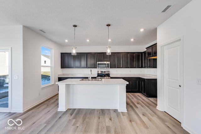 kitchen with sink, hanging light fixtures, stainless steel appliances, an island with sink, and light wood-type flooring