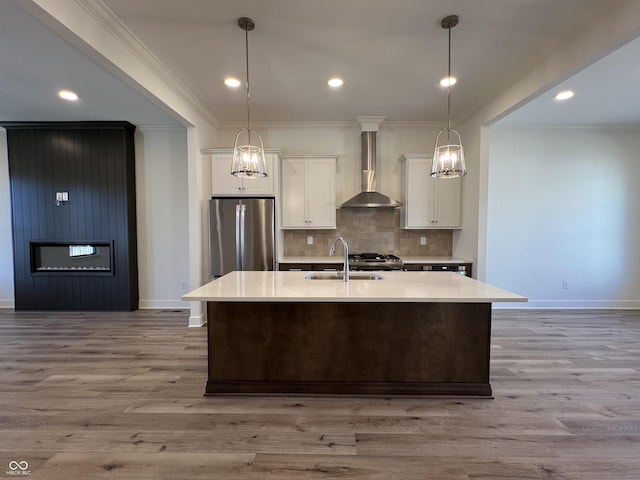 kitchen featuring an island with sink, stainless steel appliances, white cabinetry, wall chimney exhaust hood, and a sink