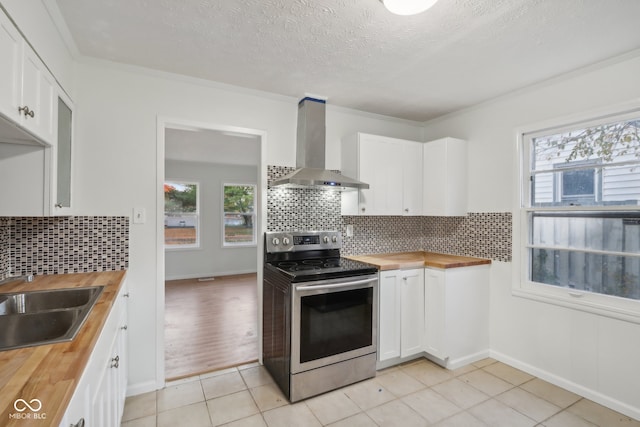 kitchen with a healthy amount of sunlight, stainless steel range with electric stovetop, white cabinetry, wall chimney exhaust hood, and wooden counters