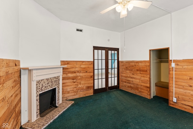 unfurnished living room featuring a tile fireplace, dark carpet, ceiling fan, french doors, and wooden walls