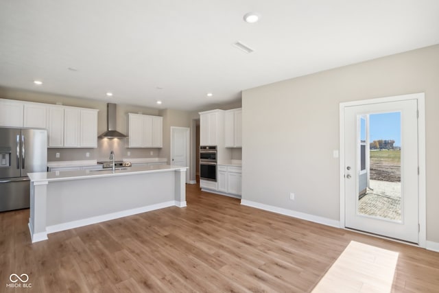 kitchen featuring appliances with stainless steel finishes, light wood-type flooring, wall chimney exhaust hood, a center island with sink, and white cabinetry