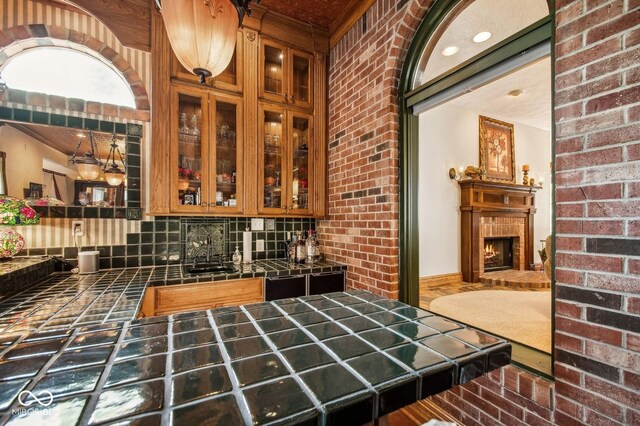 6902 Creekridge Trl, Indianapolis, IN 46256 - kitchen featuring tile countertops, decorative backsplash, brick wall, glass insert cabinets, and a lit fireplace