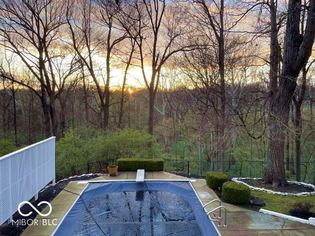 pool at dusk with a diving board, fence, a covered pool, and a view of trees