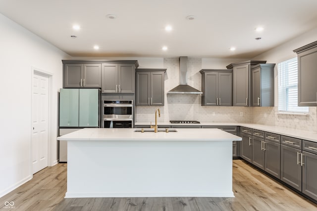 kitchen featuring sink, appliances with stainless steel finishes, wall chimney exhaust hood, a kitchen island with sink, and light wood-type flooring