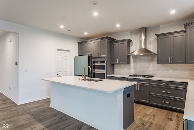 kitchen with light wood-type flooring, wall chimney range hood, appliances with stainless steel finishes, and a kitchen island with sink