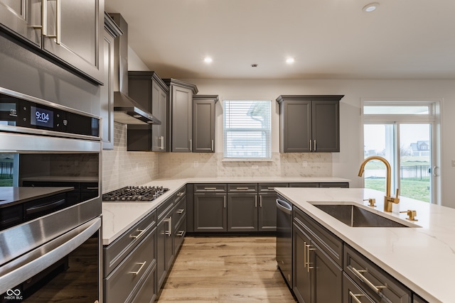 kitchen featuring wall chimney range hood, appliances with stainless steel finishes, decorative backsplash, sink, and light hardwood / wood-style flooring