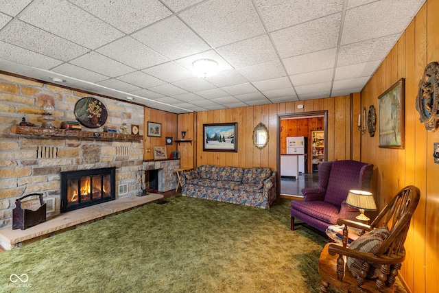 living room featuring a drop ceiling, a stone fireplace, carpet floors, and wooden walls