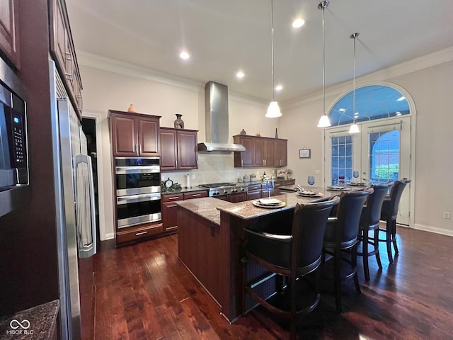 kitchen with wall chimney range hood, appliances with stainless steel finishes, dark hardwood / wood-style floors, pendant lighting, and a kitchen island with sink