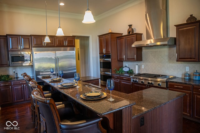 kitchen with wall chimney range hood, a large island, built in appliances, dark hardwood / wood-style floors, and decorative backsplash