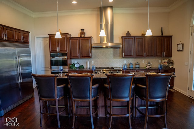 kitchen with a center island with sink, appliances with stainless steel finishes, wall chimney exhaust hood, hanging light fixtures, and dark hardwood / wood-style flooring