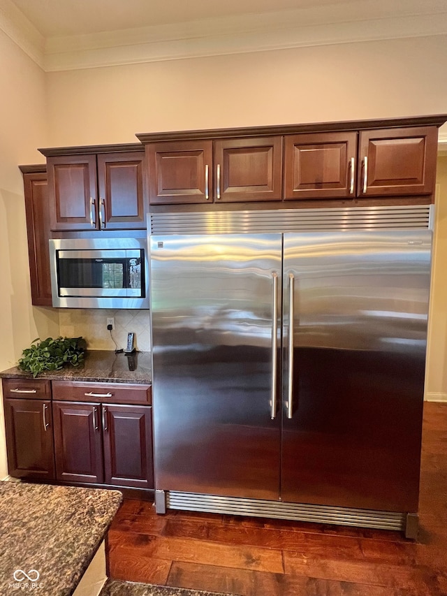 kitchen featuring built in appliances, tasteful backsplash, ornamental molding, dark hardwood / wood-style floors, and dark stone countertops