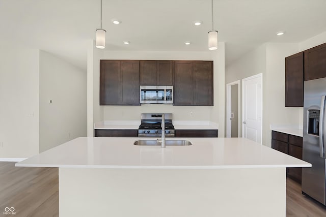 kitchen featuring appliances with stainless steel finishes, decorative light fixtures, dark brown cabinets, light hardwood / wood-style floors, and a kitchen island with sink