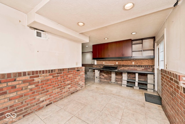 kitchen with a textured ceiling, dark brown cabinets, light tile patterned flooring, and brick wall