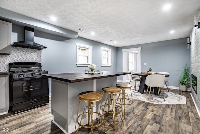 kitchen with gas stove, wall chimney exhaust hood, butcher block countertops, a breakfast bar area, and hardwood / wood-style flooring