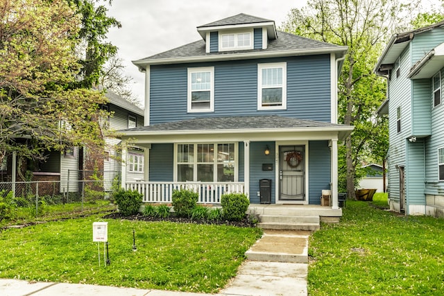 american foursquare style home with a front yard, a shingled roof, covered porch, and fence