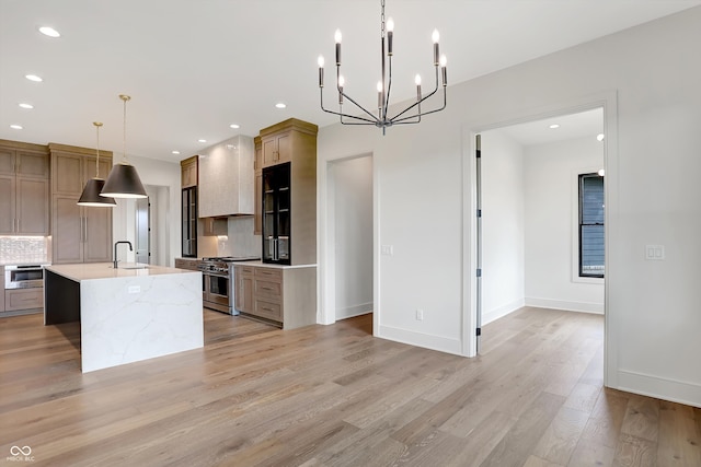 kitchen featuring premium range hood, backsplash, stainless steel stove, decorative light fixtures, and an island with sink