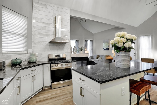 kitchen with wall chimney exhaust hood, stainless steel gas range oven, dark stone counters, vaulted ceiling, and light wood-type flooring