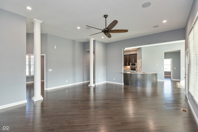 unfurnished living room with ornate columns, a wealth of natural light, dark wood-type flooring, and ceiling fan