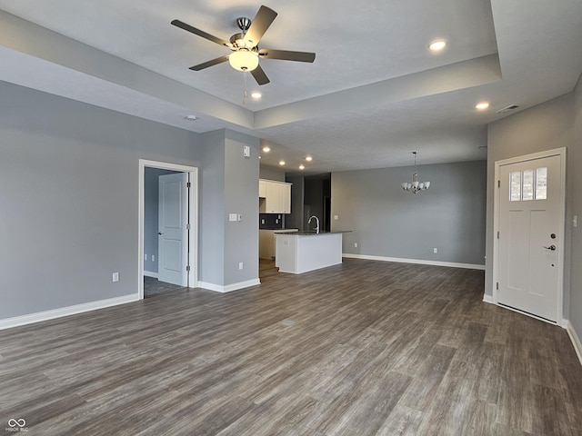 unfurnished living room featuring sink, ceiling fan with notable chandelier, dark wood-type flooring, and a tray ceiling