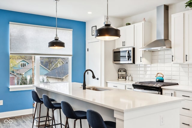 kitchen featuring stainless steel appliances, sink, wall chimney range hood, pendant lighting, and a center island with sink