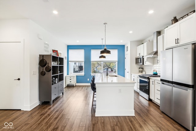 kitchen with appliances with stainless steel finishes, wall chimney exhaust hood, dark wood-type flooring, white cabinetry, and an island with sink