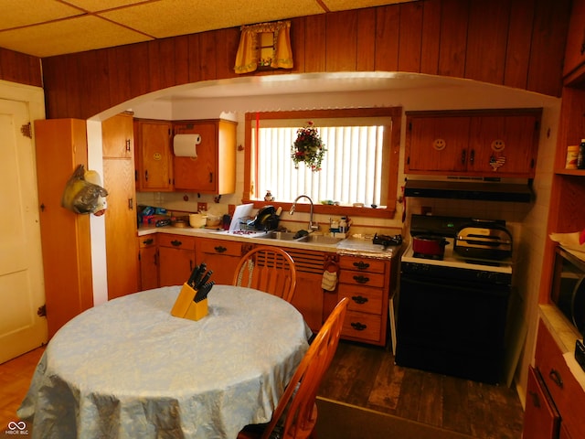 kitchen with a drop ceiling, black range with gas stovetop, sink, wooden walls, and dark hardwood / wood-style floors