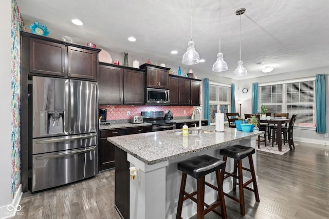 kitchen featuring a kitchen island with sink, hanging light fixtures, appliances with stainless steel finishes, dark brown cabinets, and dark hardwood / wood-style flooring